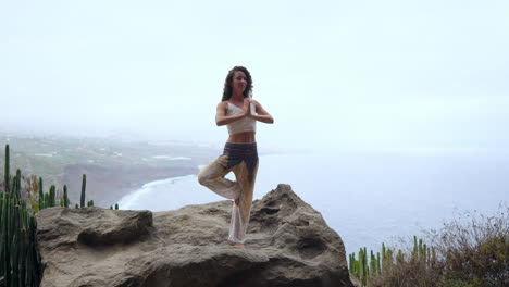 On-an-island-amidst-mountains,-a-young-woman-practices-yoga,-standing-on-one-leg,-arms-raised,-overlooking-the-ocean